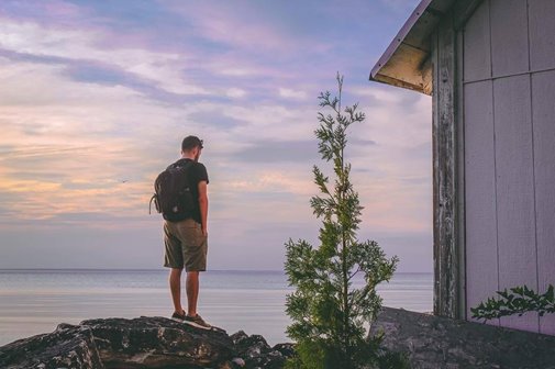 A man standing on a rock next to a wooden building looking out onto the lake.