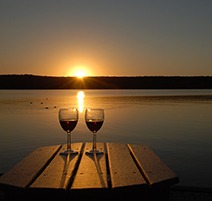 Two glasses of wine sitting on a table in front of the sun setting over the lake.