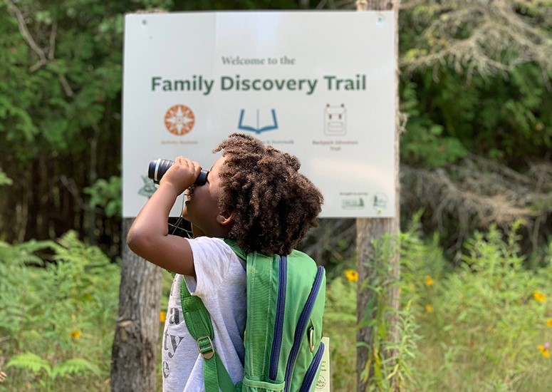 A little kid looking into binoculars near the Family Discovery Trail sign.