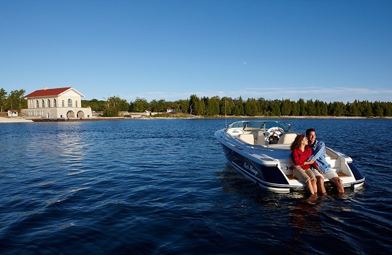 Two people with there feet in the lake sitting on the back of a boat with a stone boathouse in the distance.