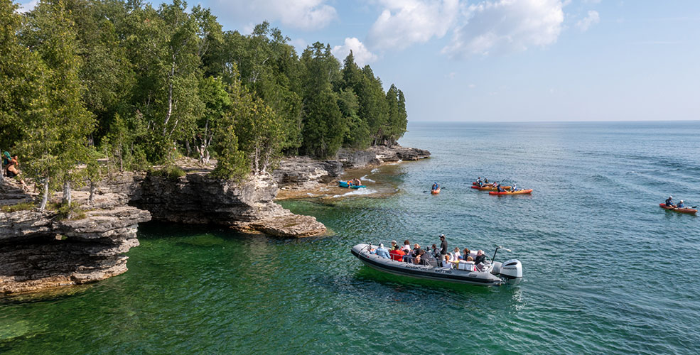 People in a boat pulling up with a tree-lined cliff line.