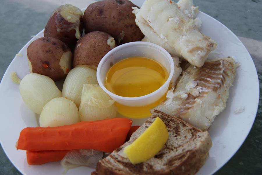 A plate full of boiled fish, potatoes, vegetables, drawn butter, and bread.