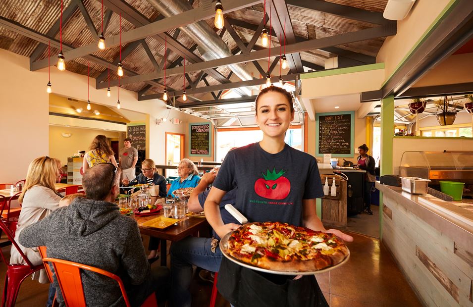 Woman showing a pizza to the camera next to a table full of diners in a restaurant.