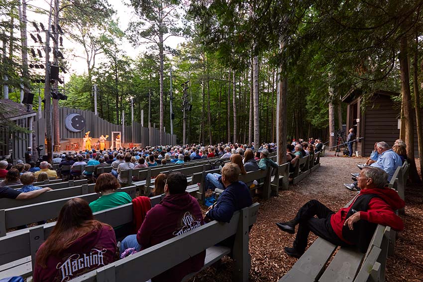 An audience watches a play at an outdoor theater in the forest.