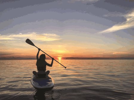 A person sitting on a paddle board with the paddle raised and the sun setting in the background