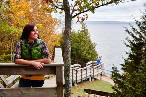 A woman stands atop a wooden staircase among fall leaves, leading to a dramatic view of Lake Michigan.