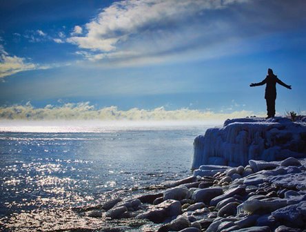 The silhouette of a person with their arms outstretched standing on a snowy rock at the lakefront.