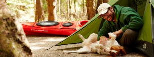 Man playing with a dog in front of a tent with a kayak in the background.