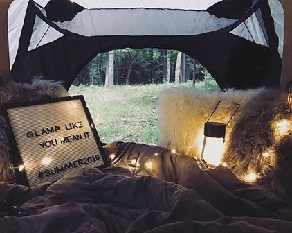 A tent with furry pillows and a comforter in front of a sign that says Glamp Like You Mean It.