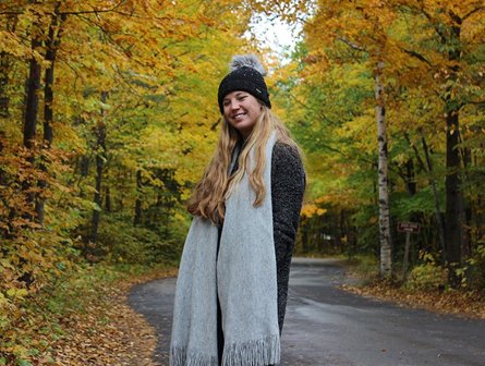 A woman with a hat and scarf standing in front of trees in their fall colors.
