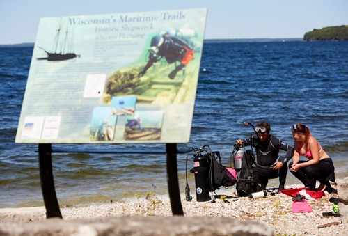Two scuba divers prep for a dive beside Lake Michigan and a colorful informational sign.