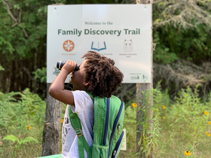 A young girl looks toward the sky with binoculars in front a trailhead sign