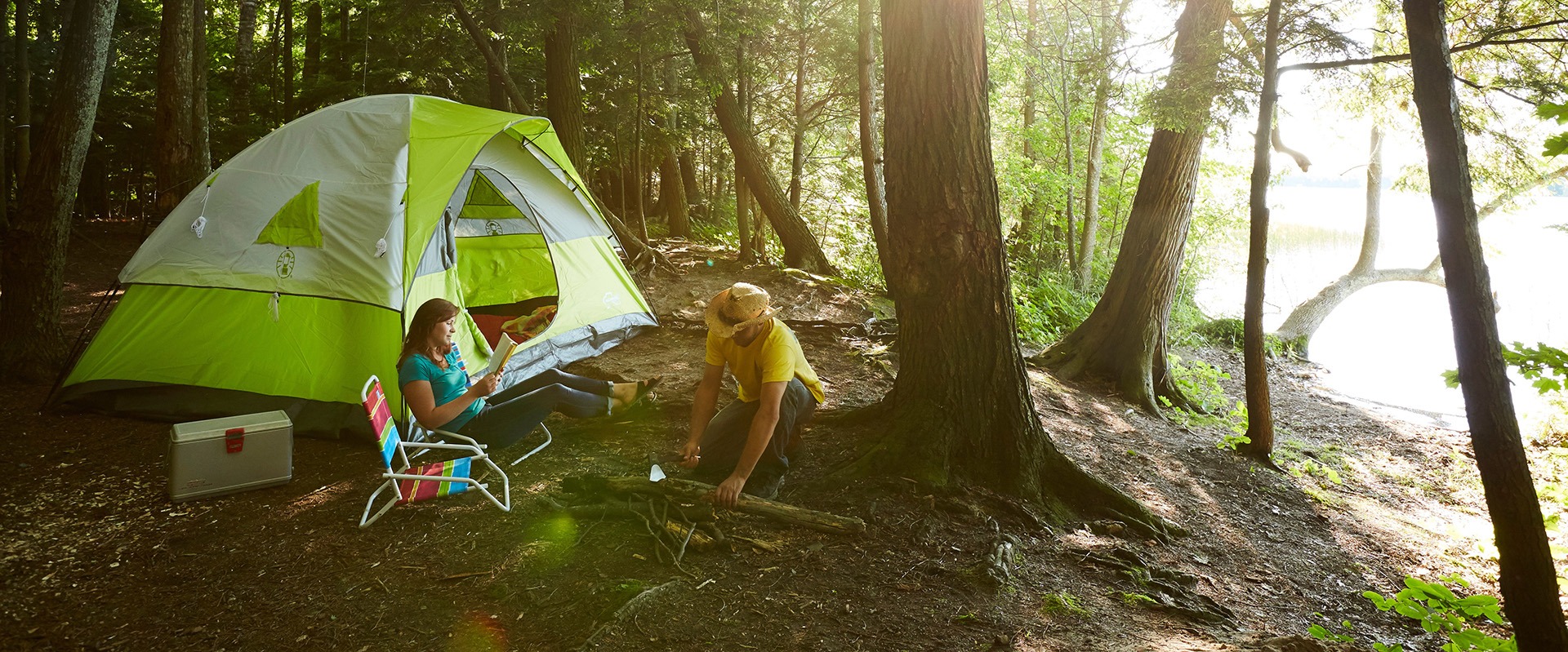 A couple does camping activities near their tent in the woods, by a lake.