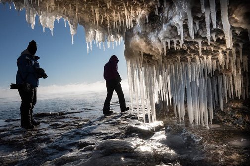 Two people walking just outside an ice-filled cave at the lakefront.