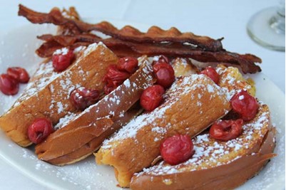 A beautiful plate of french toast covered in cherries and powdered sugar and accompanied by bacon.