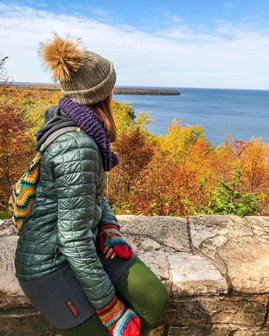 A woman bundled up sitting on a stone wall looking out at colorful trees and the lake.