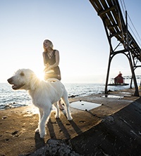 A woman walking a dog on a pier with a lighthouse in the distance.