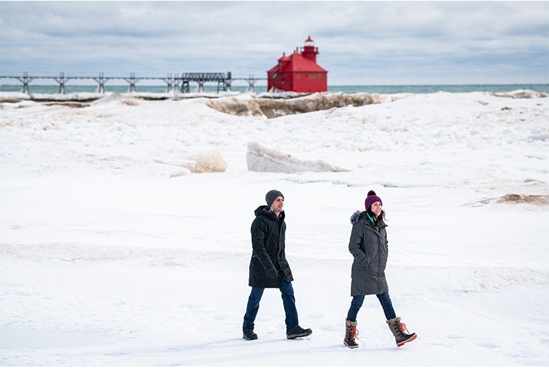 Two people walking through the snow with a red lighthouse in the distance