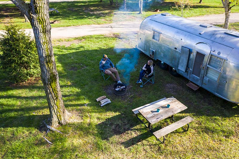 A couple sitting outside of their airstream camper next to a camp fire