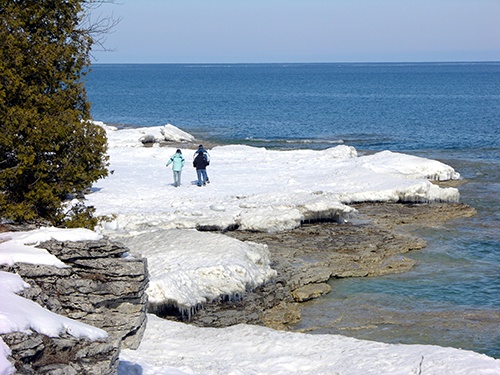 Two people hike on snow-covered rocks at Cave Point.