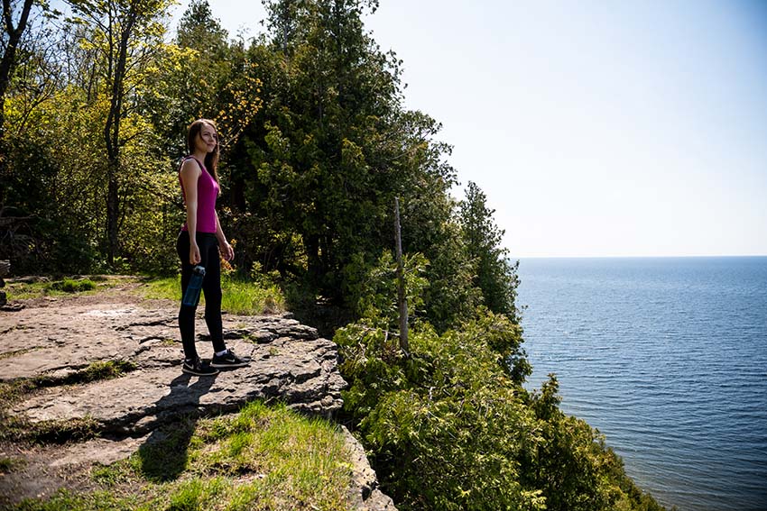 A woman stands atop a massive forested bluff, looking out over the lake.