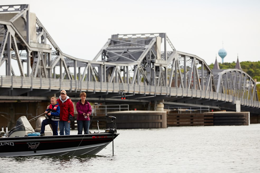 A family fishing off of a boat with a steel bridge in the background.