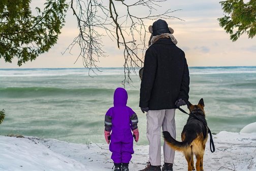 A person with a child and a dog walking through the snow at the lakefront.