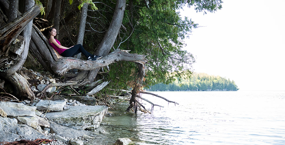 A woman sitting on a low tree branch along the lake.