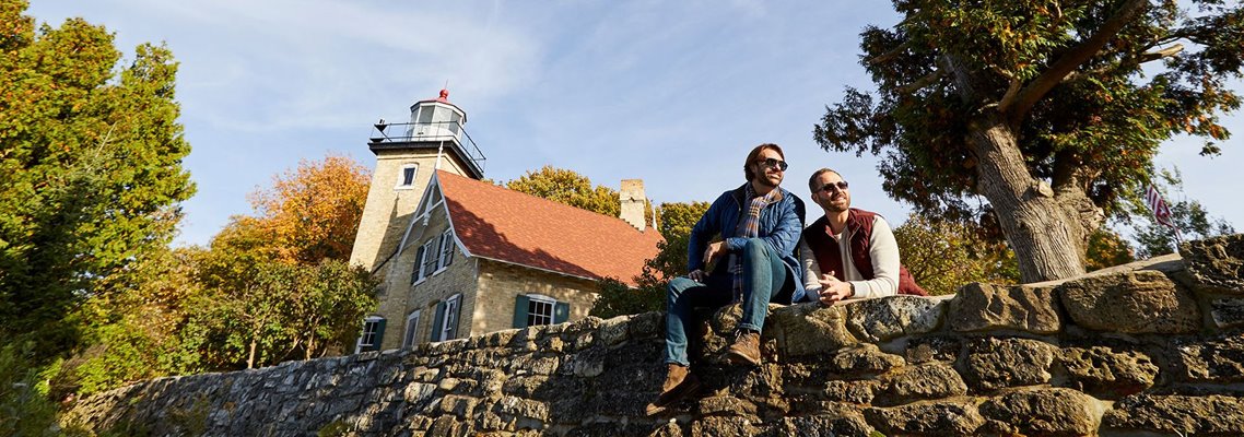 A couple sitting on a stone wall in front of a lighthouse.