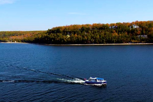 A boat tour glides across deep-blue water with a fall-colored shoreline in the background.