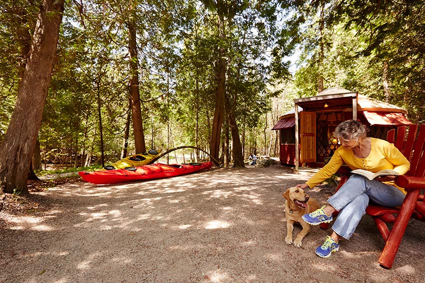 A woman and her dog sitting outside a cabin with a kayak in the background.