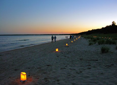 Paper bag lanterns lighting up the sandy beach along the lake shoreline with people walking by