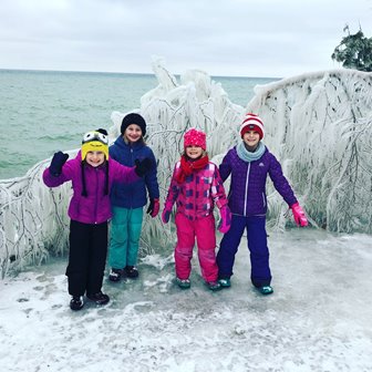 Children bundled up and smiling in front of ice-covered trees at the lakefront.
