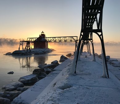 The sun rising beyond a lighthouse at the end of a snow-covered pier.