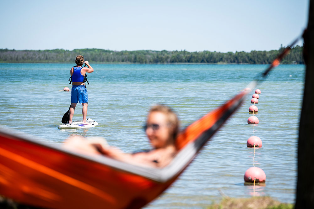Woman in a hammock near the lake with someone paddle boarding in the background