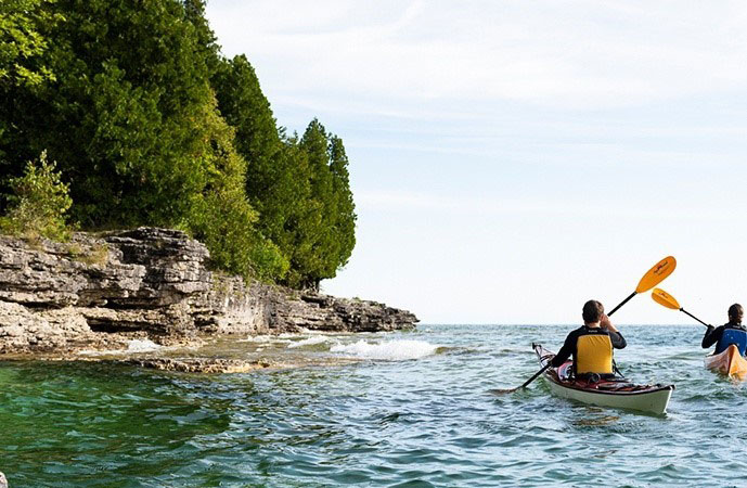Two people kayaking close to cliff-lined shore.
