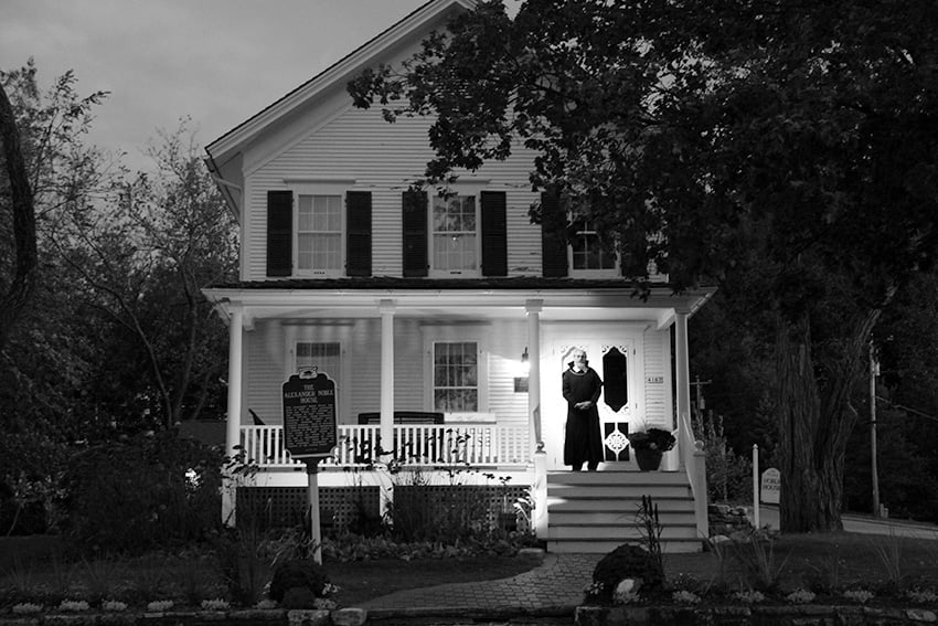 An eerie man stands on the porch of the old, haunted Noble House.
