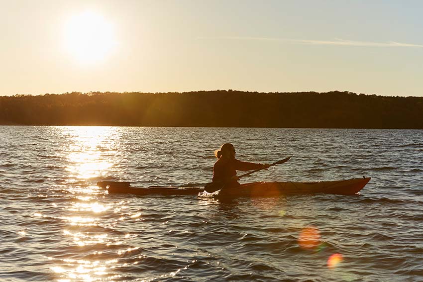 A man kayaks on a lake with a forested shoreline at sunset.